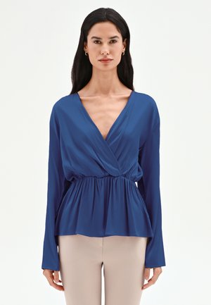 Femme aux longs cheveux foncés portant un chemisier à manches longues bleu profond avec un lien et un pantalon beige clair, debout devant un fond blanc uni.