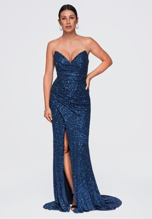 Femme portant une robe bustier bleu foncé pailletée avec une fente haute sur la cuisse et des boucles d'oreilles en forme d'anneau, debout devant un fond clair uni.