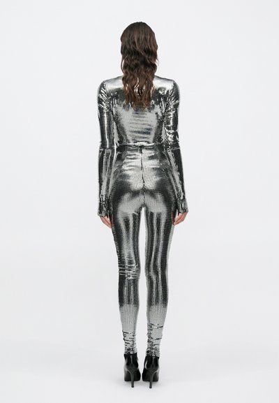 Sieviete stāv ar muguru pret skatītāju, tērpusies sudraba metāliskā garām piedurknēm cieši piegulošā kombinezonā un melnos augstpapēžu apavos uz balta fona.