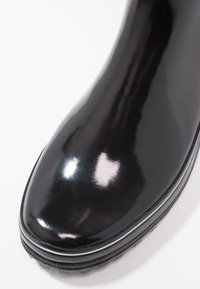 Botte en caoutchouc noir poli avec un bout arrondi, une semelle texturée et un subtil accent blanc à la base. Finition lisse et brillante.