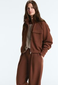 Ženska, ki nosi rjavo jakno z zadrgo in zavihki na žepih preko rjavo-bele črtaste majice ter ujemajoče se rjave široke hlače.