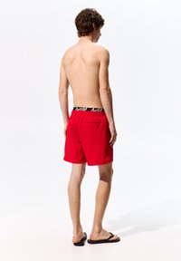 Pantaloni da bagno rossi con una vita elastica. Include una tasca sul retro con cerniera. Il materiale appare leggero e ad asciugatura rapida.