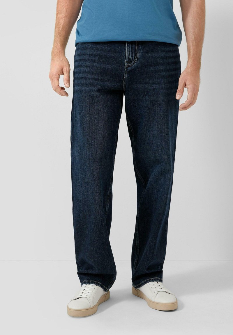 Niebieskie jeansy z ciemnym praniem o prostych nogawkach, z przeszyciami i kieszeniami, zestawione z białymi trampkami.