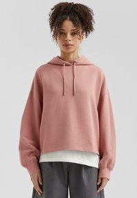 Roze gestructureerde hoodie met een trekkoordkap, verlaagde schouders en ribgebreide manchetten, gelaagd over een wit shirt en gecombineerd met grijze broeken.
