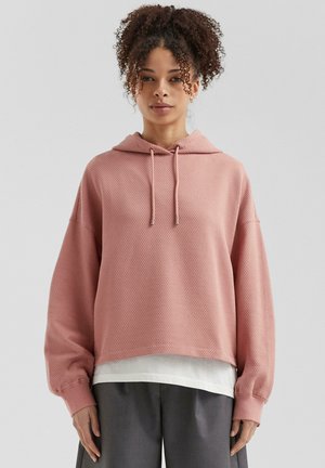 Roze gestructureerde hoodie met een trekkoordkap, verlaagde schouders en ribgebreide manchetten, gelaagd over een wit shirt en gecombineerd met grijze broeken.