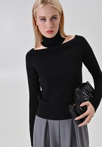 Top nero a maglia con design off-shoulder e colletto alto. Gonna grigia plissettata. Borsa nera con dettagli in argento e diversi scomparti.