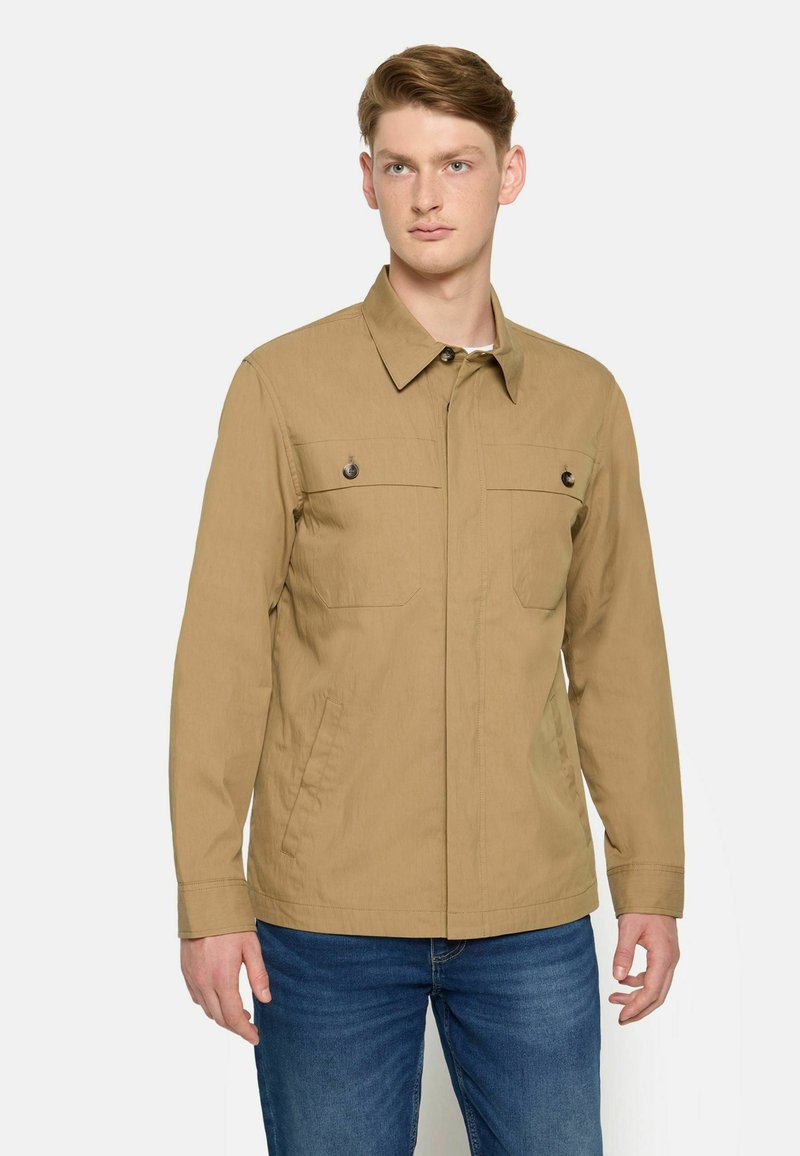 Lys tan jakke lavet af et tekstureret stof, med en knap-ned krave, to brystlommer med knapper og lange ærmer. Bæres sammen med denim jeans.