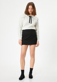 Weißer gerippter Pullover mit dekorativen Schulterdetails und schwarzem Schleifenband, kombiniert mit einem taillierten schwarzen Minirock. Schwarze knöchellange Socken und Ballerinas.