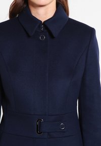 Manteau bleu marine en tissu texturé, avec un col pointu, une fermeture éclair et une taille ceinturée ornée d'un accent métallique circulaire.