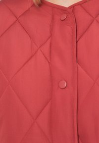 Rød quilted jakke med diamantsting og to runde trykknapper langs midtforkjæringen.