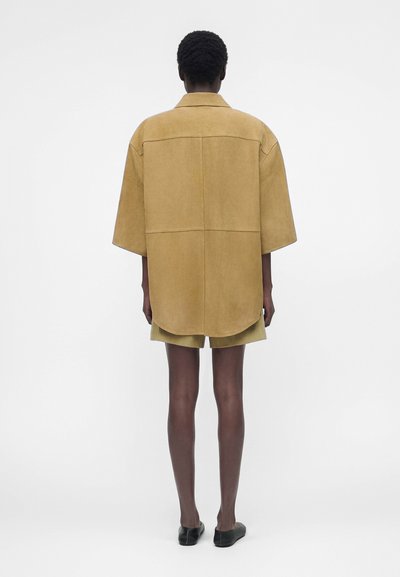 Άτομο με σκουρόχρωμο δέρμα και κοντά μαλλιά στέκεται με την πλάτη γυρισμένη, φοράει άνετο μπεζ κοντομάνικο πουκάμισο, ταιριαστό σορτσάκι και μαύρα παπούτσια σε λευκό φόντο.