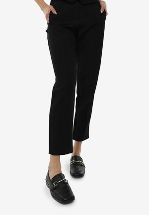Schwarze Loafer mit strukturiertem Finish, ergänzt durch eine goldfarbene Kettendetails. Kombiniert mit maßgeschneiderten schwarzen Hosen im schmalen Schnitt.
