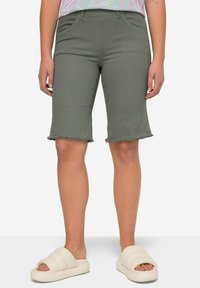 Pantalones cortos de color verde oliva, hasta la rodilla, en una tela suave y elástica con un dobladillo deshilachado. Combinados con sandalias chunky, blancas, tipo slip-on.