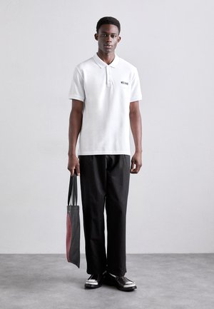 White polo shirt with short sleeves, black pants, and black shoes. The model holds a black bag with a maroon accent. Simple and clean design.