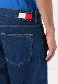 Donkerblauwe denim jeans met zichtbare stiksels, voorzien van een Tommy Jeans logo patch in marineblauw, wit en rood op de achterste tailleband.