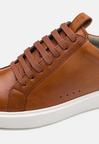 Brune lær-sneaker med rund tå, teksturert overflate, flate lisser og hvit gummisåle. Har perforeringer for ventilasjon.