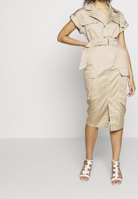 Abito utility beige in cotone, con maniche corte arrotolate, tasche frontali, vita con cintura e orlo lungo il ginocchio con spacco.