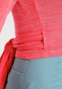 Korálový pletený top s detailem vázání v pase, kombinovaný s texturovanou světle modrou žebrovanou sukní. Materiál vypadá měkce a elasticky.