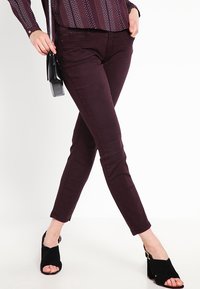 Bordó úzké džíny z měkkého denimu, s elegantním designem a jemnými detaily švů, spárované s černými sandály na podpatku.