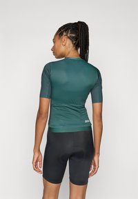 Tricou de ciclism cu mânecă scurtă în verde închis, cu un design ajustat, textură netedă și detalii de buzunar la spate, asociat cu pantaloni scurți negri de ciclism.
