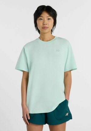 Jonge vrouw met kort zwart haar, die een lichtgroen oversized T-shirt en donkergroene sportkorte broek draagt tegen een effen achtergrond.
