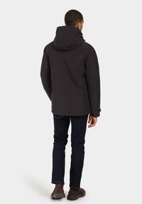 Črna puhovka z odstranljivo kapuco, elegantnega dizajna in elastičnimi manšetami, v kombinaciji z temno modrimi kavbojkami in rjavimi čevlji.