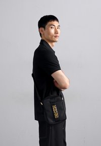 Geantă de umăr neagră dintr-un material texturat, cu un logo auriu "Moschino", formă rectangulară și curea reglabilă.