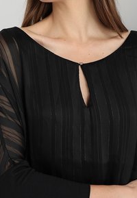 Blusa nera con righe verticali e scollatura a chiave. Il tessuto trasparente e motivo sulle maniche aggiunge texture e dettagli.