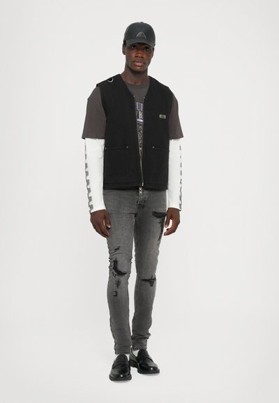 Vest negru fără mâneci, cu două buzunare frontale, purtat peste un tricou grafic închis și manșete albe lungi. Blugi gri distruși și pantofi negri completează look-ul.