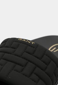 Μαύρο σανδάλι με σαγιονάρα και επενδυμένο, πλεγμένο λουράκι. Διαθέτει χρυσό λογότυπο "GANT" και λεία σόλα. Τονίζει την άνεση και το μοντέρνο σχεδιασμό.