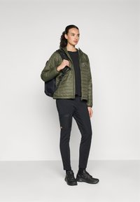 Lichte, olijfgroene gewatteerde jas met rits, zwarte sweatshirt, zwarte cargobroek en zwarte sneakers. Model houdt een zwarte rugzak vast.