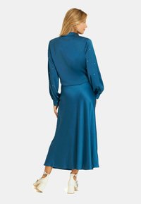 Vestido de satén azul verdoso con mangas largas, hombros plisados y adornos en las mangas, combinado con zapatos de tacón cuadrado blancos. Vista desde atrás.