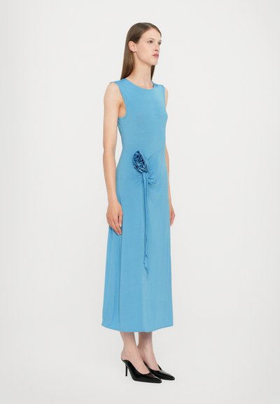 Rochie fără mâneci, de culoare albastru deschis, cu un bust ajustat, detaliu drapat pe lateral cu un accent floral și fustă lungime ankle. Poartă tocuri negre.