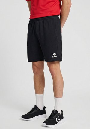 Mand iført sorte hummel-shorts, hvide ankelsokker, sorte og hvide sneakers og en rød skjorte, stående mod en ensfarvet baggrund.