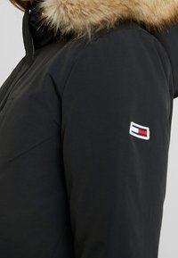 Black padded jacket featuring a removable fur collar and a small logo patch with red, white, and blue elements on the sleeve.