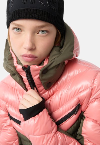 Puffjacke mit pinken und olivgrünen Einsätzen, hoher Kragen, mit Reißverschlusstaschen und einer schwarzen Mütze mit einem Logo auf der Vorderseite.