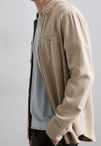 Beige nappisomisteinen kauluspaita, jossa rintatasku, kerroksittain vaaleansinisen t-paidan päällä. Kangas vaikuttaa pehmeältä, rento istuvuus ja hienovarainen tekstuuri.