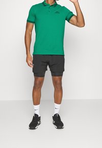 Zielona koszulka polo z czarnymi paskami przy kołnierzu i rękawach, zestawiona z czarnymi szortami i czarnymi butami sportowymi. Białe skarpetki z logo.