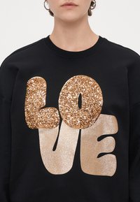 Μαύρη μπλούζα με χρυσά γράμματα από γκλίτερ που γράφουν "ΑΓΑΠΗ". Λείο υλικό με λαμπερες λεπτομέρειες σε ένα χαλαρό σχέδιο.