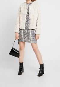 Giacca imbottita color crema con finitura testurizzata, indossata sopra un vestito aderente con motivo a pelle di serpente. Stivaletti neri con tacco largo, che tengono una piccola borsa nera.
