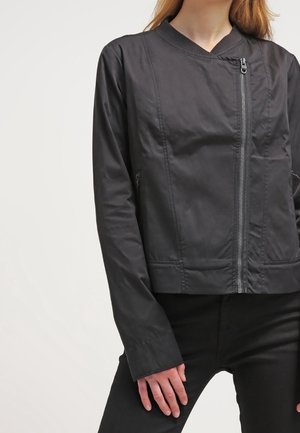 Frau trägt eine schwarze Jacke mit diagonalem Frontreißverschluss, kombiniert mit schwarzen Hosen, vor weißem Hintergrund.