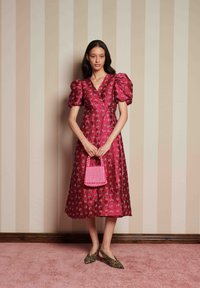 Vestido de seda com padrão floral rosa e mangas bufantes, combinado com uma pequena bolsa xadrez rosa e sapatos de salto de estampa de leopardo, posando contra um fundo listrado.