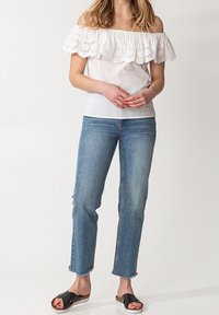 Top bianco scollato sulle spalle con dettaglio arricciato in pizzo, abbinato a jeans dritti blu chiaro e sandali neri con suole bianche.