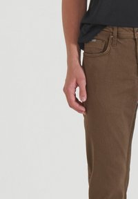 Los pantalones de mezclilla marrones presentan un ajuste slim con bolsillos frontales y una cinturilla visible. El tejido tiene una textura suave con detalles de costura sutiles.