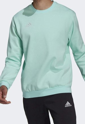 Mand iført lyseblå Adidas sweatshirt og sorte Adidas bukser, står med armene let udstrakte.
