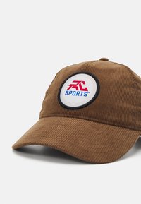 Pruunist sametist müts, millel on ümmargune patch punase ja sinise "RC SPORTS" logoga, rõhutades tekstuuri ja disainielemente.