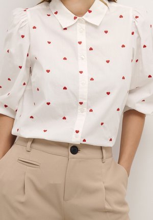 Witte blouse met knopen en rode hartpatronen, korte pofmouwen, gecombineerd met beige broek met hoge taille en een zwarte knoop.