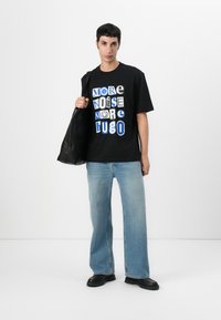 Svart bomull t-skjorte med hvit og blå grafisk tekst «MER STØY MER HUGO», sammen med lyseblå løse jeans og svarte sko.