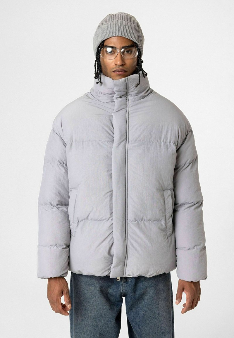 Grijze puffer jacket met hoge kraag, ritssluiting en gequilt textuur; gecombineerd met een grijze geribde beanie en een doorschijnende bril. Casual fit.