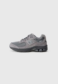 Pelēki sporta apavi ar strukturētu sieta un ādas augšdaļu; tiem ir mīkstināta zole un izcila New Balance logotipa zīme sānos.